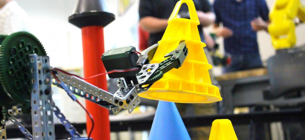 Student-built and programed VEX robot moving a cone with students in the background