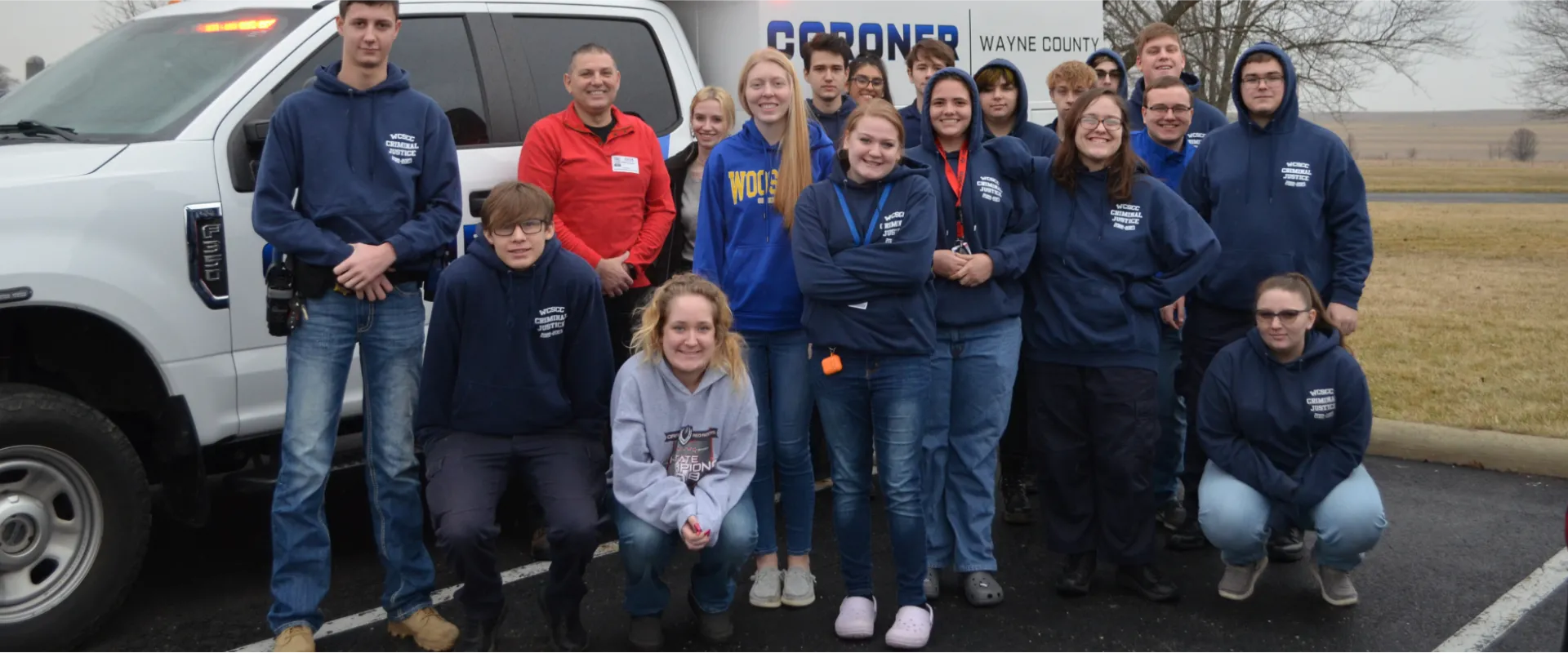 A group of people wearing matching navy hoodies pose in front of a white vehicle labeled "Coroner" in a parking lot with fields and trees in the background.