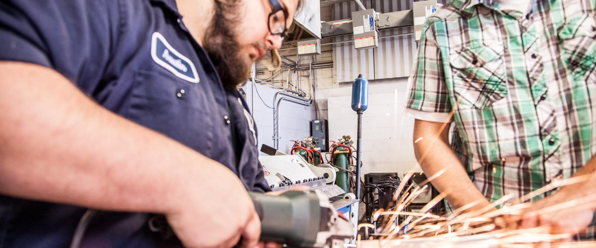 Two men, one is using a grinder to cut metal and the other is observing. 