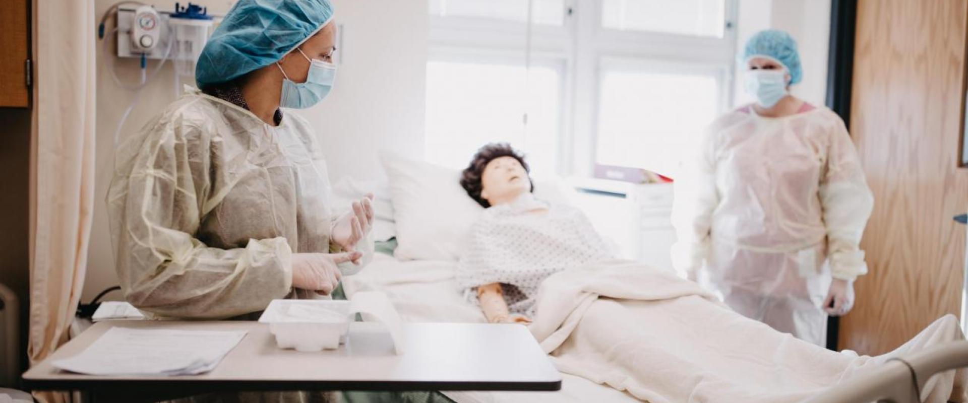 two nurses who are wearing hair covers, masks, gloves and splatter coats on either side of a hospital bed where a test dummy lies