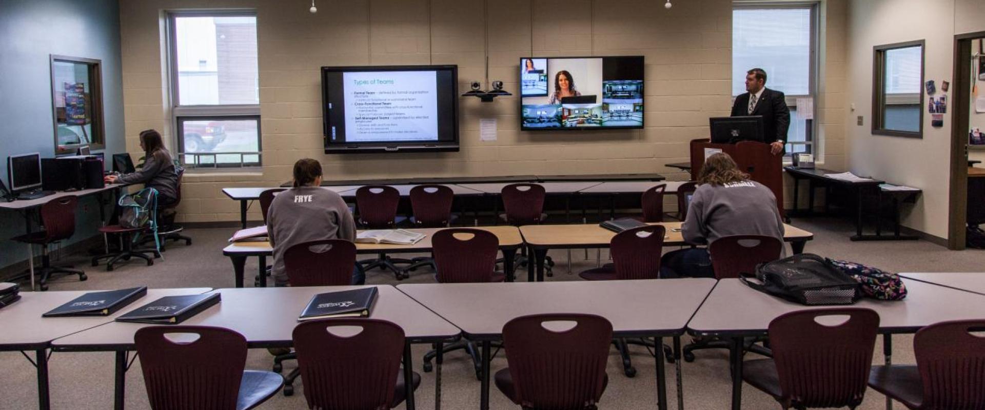 Three students in a class room while there is a teacher at a podium