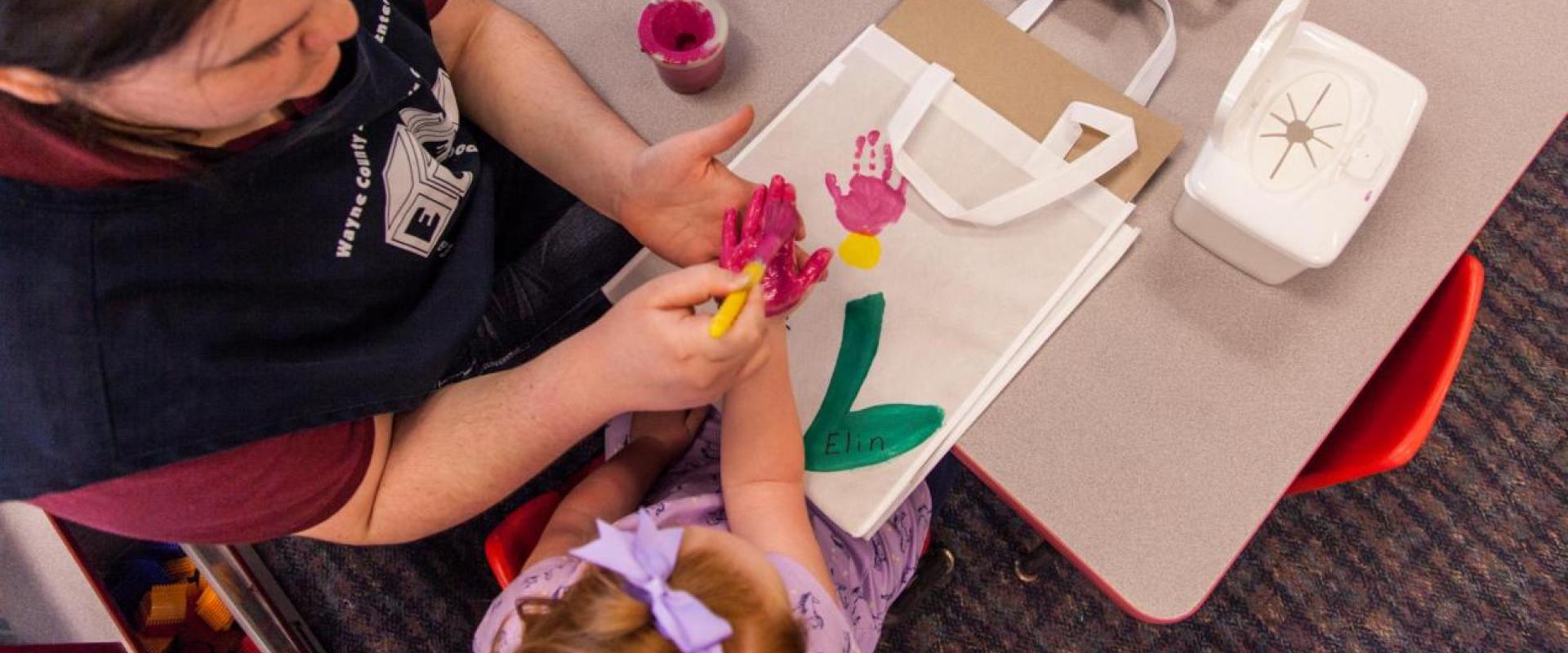 A daycare teacher assisting a young girl with an activity by painting her hand
