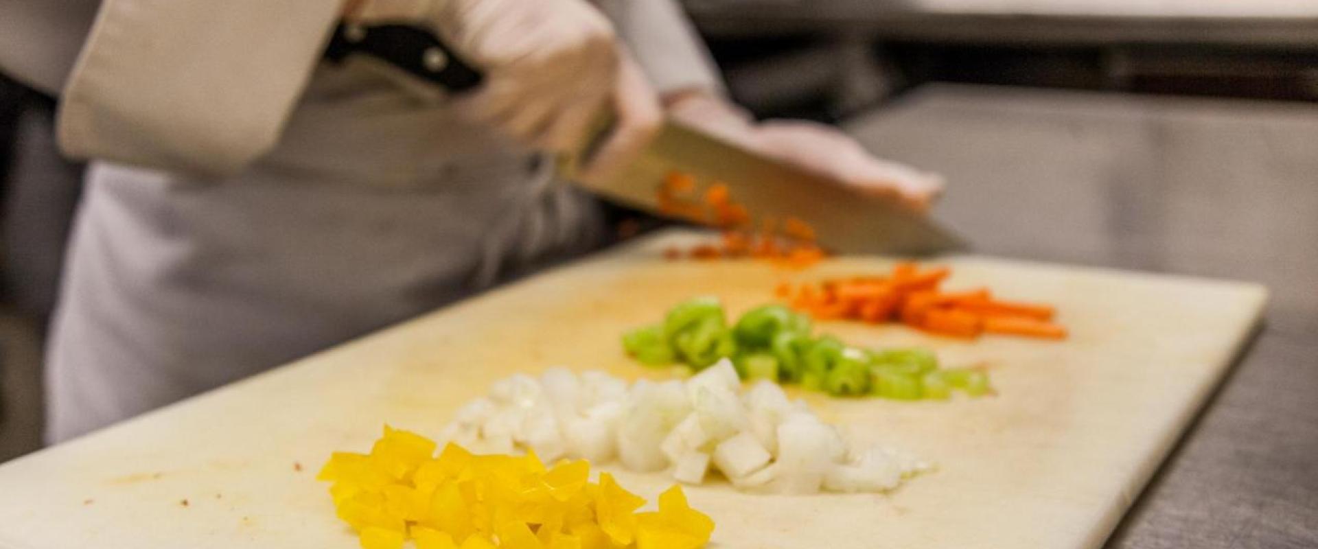 A student chopping up a red pepper. Next to it are chopped yellow peppers, onions, green peppers and carrots