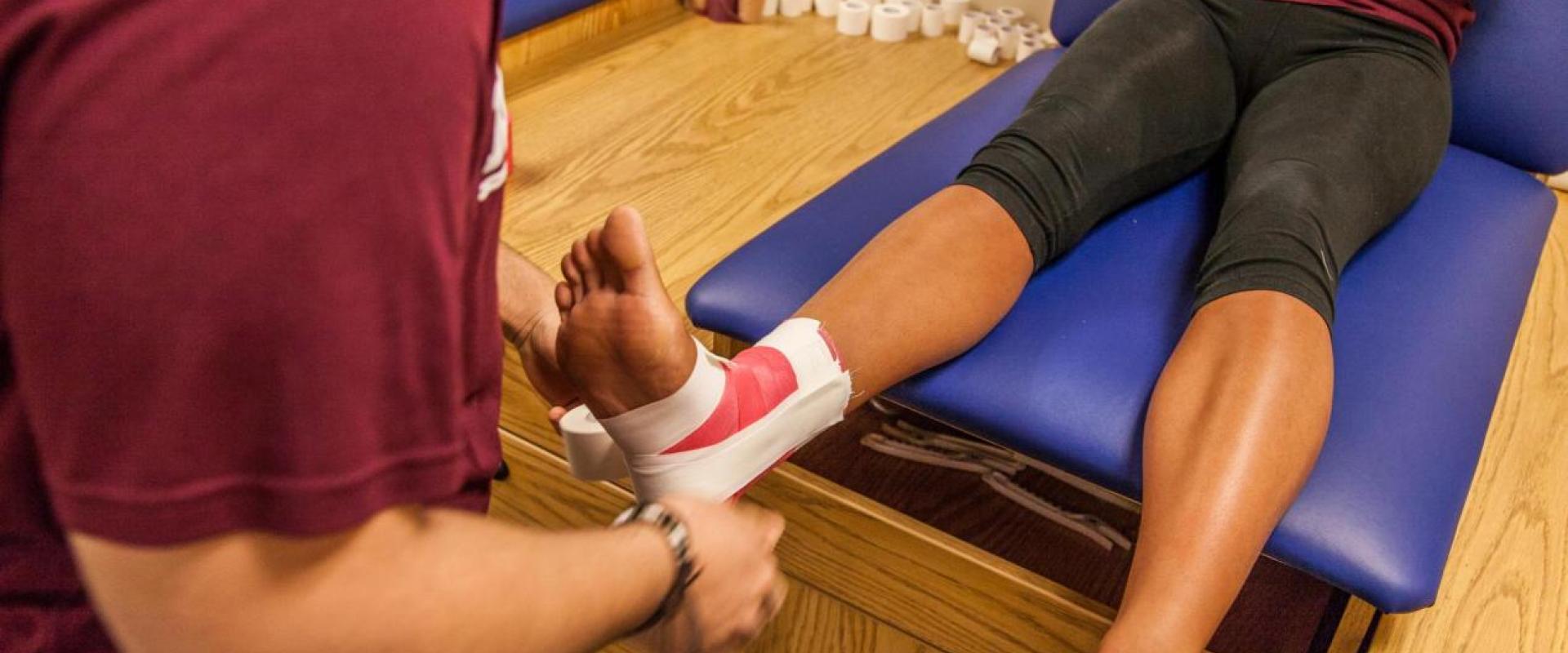 One individual wrapping another individuals ankle with athletic tape