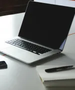 Laptop opened on a table next to a notebook with a pen.
